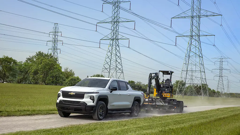 We see a white 2024 Chevy Silverado EV pickup truck towing a small front-loader. 