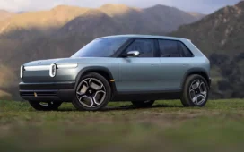 A silver Rivian R3 crossover electric utility vehicle is seen with its headlights on.